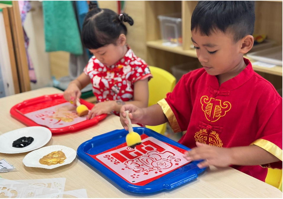 Boy and girl in red outfits paint with sponge brushes on stencil trays at a classroom table.